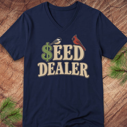 A navy blue V-neck t-shirt with a graphic reading '$EED DEALER' in beige letters, featuring a green dollar sign and two birds (a small white bird and a red cardinal), laid flat on a rustic wood background with pine branches.