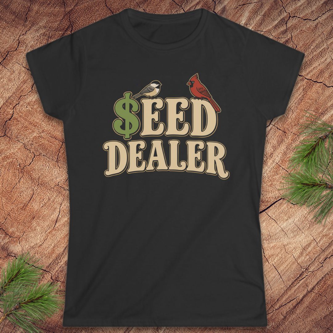 A black short-sleeve crewneck t-shirt with a retro-style graphic reading '$EED DEALER' in beige letters, featuring a green dollar sign, a small bird, and a red cardinal on a wood background with pine branches.