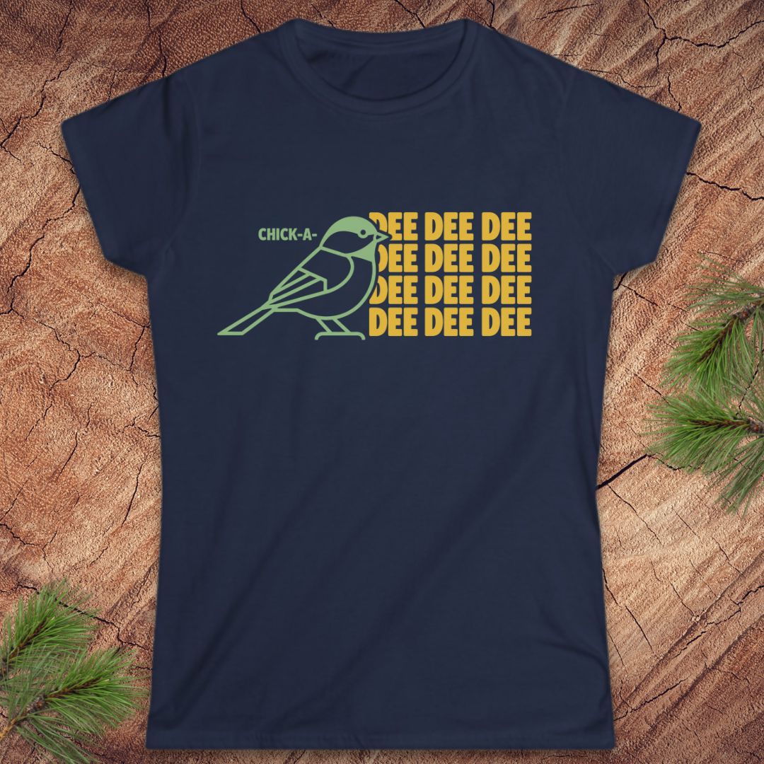 A navy blue short-sleeve t-shirt with a graphic of a green chickadee and yellow text reading 'CHICK-A- DEE DEE DEE DEE DEE DEE', laid flat on a wood surface with pine branches in the corners.