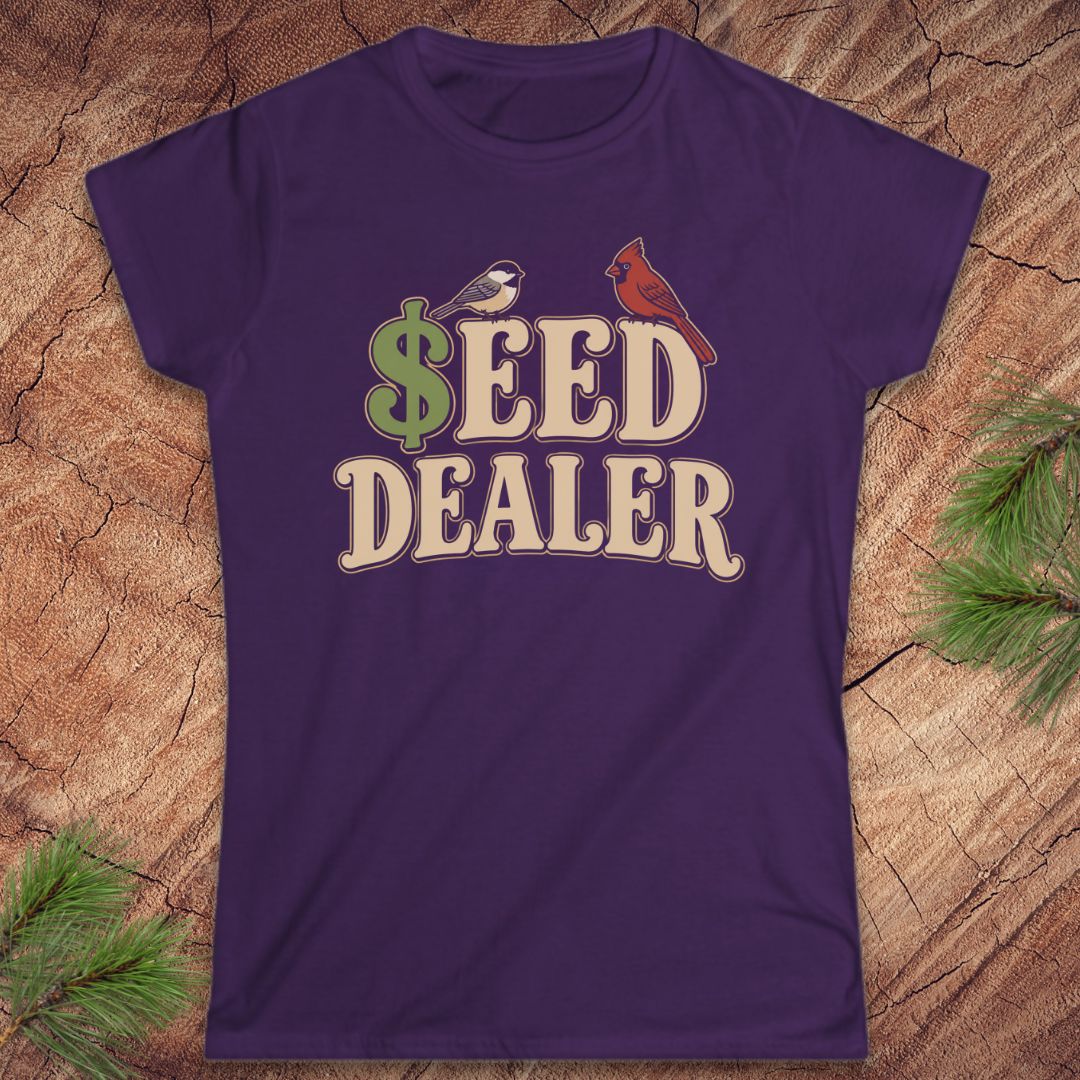 A purple short-sleeve crewneck t-shirt with a retro-style graphic reading '$EED DEALER' in beige letters, featuring a green dollar sign, a small bird, and a red cardinal on a wood background with pine branches.