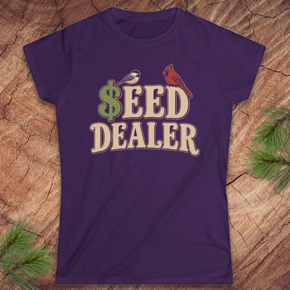 A purple short-sleeve crewneck t-shirt with a retro-style graphic reading '$EED DEALER' in beige letters, featuring a green dollar sign, a small bird, and a red cardinal on a wood background with pine branches.