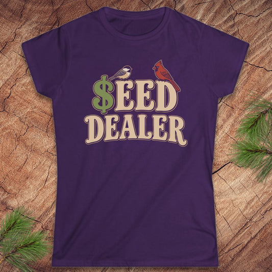 A purple short-sleeve crewneck t-shirt with a retro-style graphic reading '$EED DEALER' in beige letters, featuring a green dollar sign, a small bird, and a red cardinal on a wood background with pine branches.
