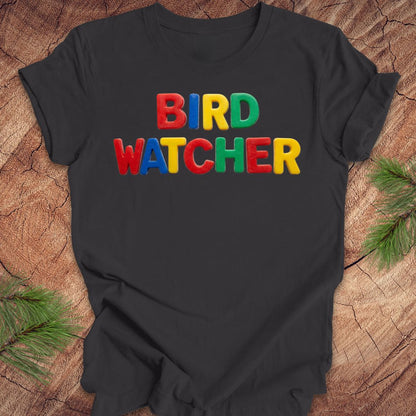 Dark gray t-shirt with 'BIRD WATCHER' in colorful letters on a wooden surface with pine branches.