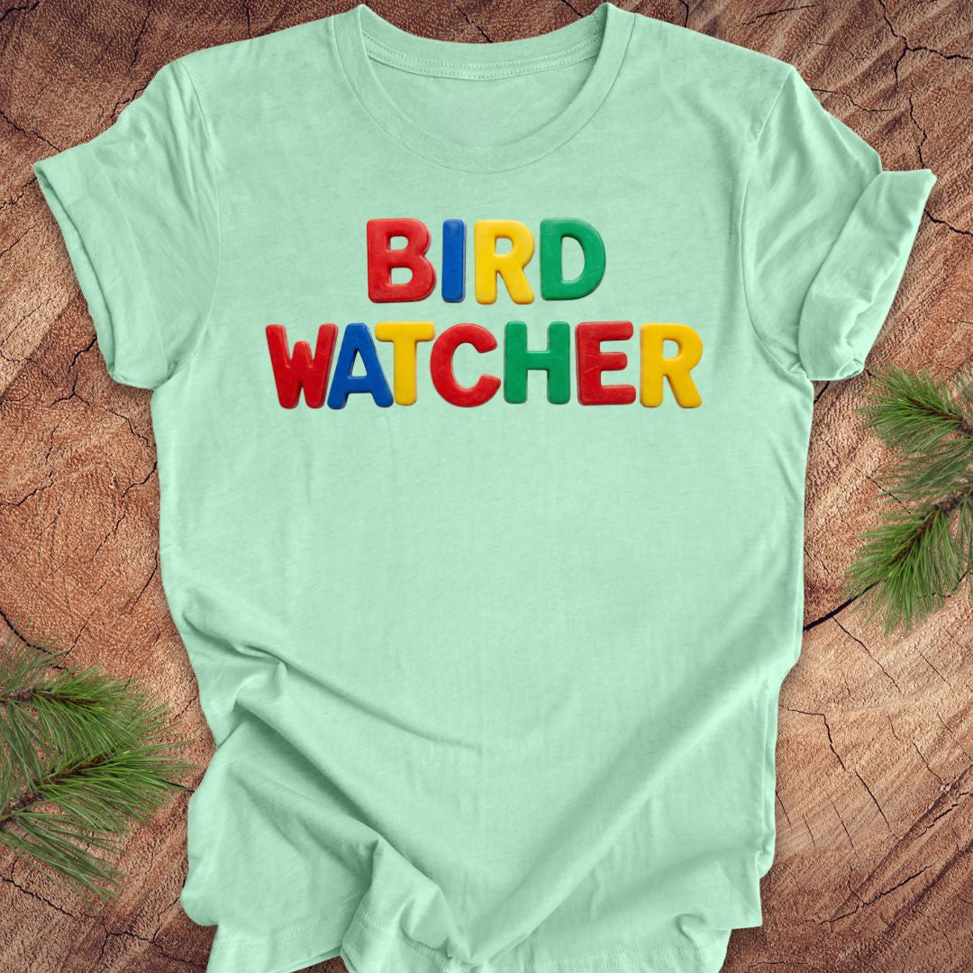 Light green t-shirt with 'BIRD WATCHER' in colorful letters on a wooden surface.