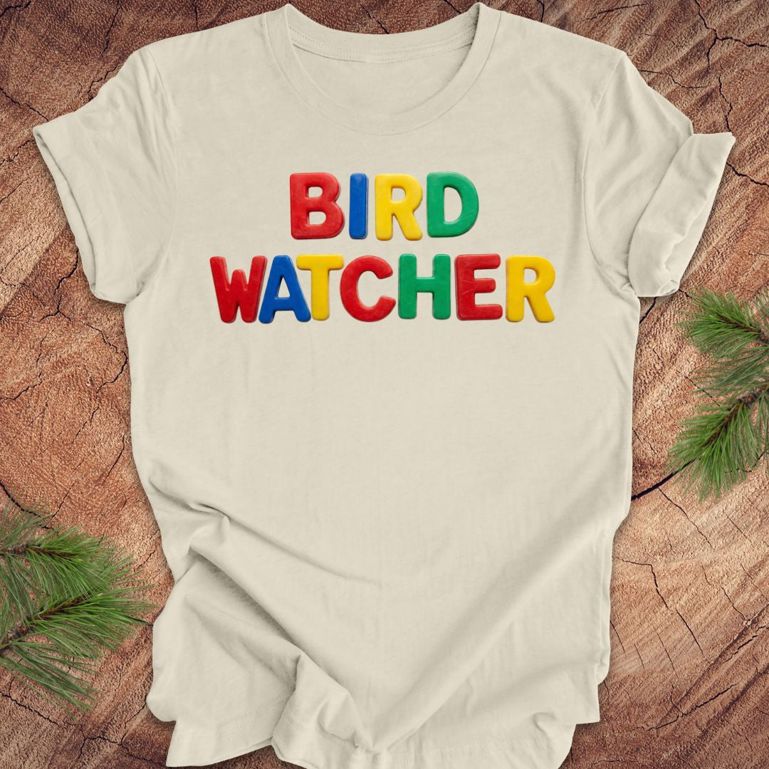 Beige t-shirt with 'BIRD WATCHER' in colorful letters on a wooden surface with greenery.