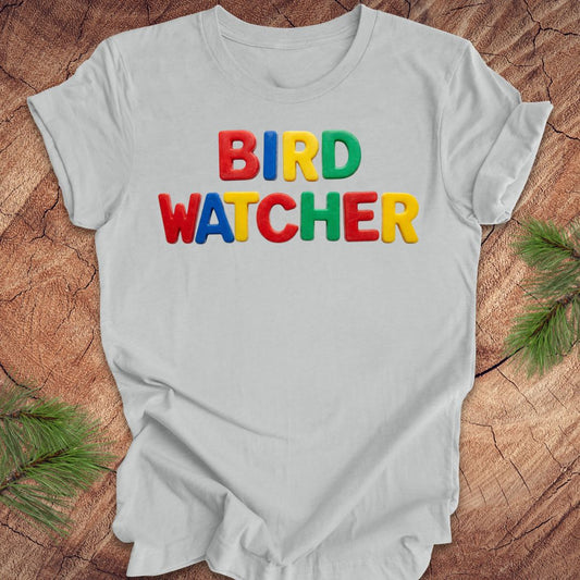Gray t-shirt with 'BIRD WATCHER' in colorful letters on a wooden surface with greenery.