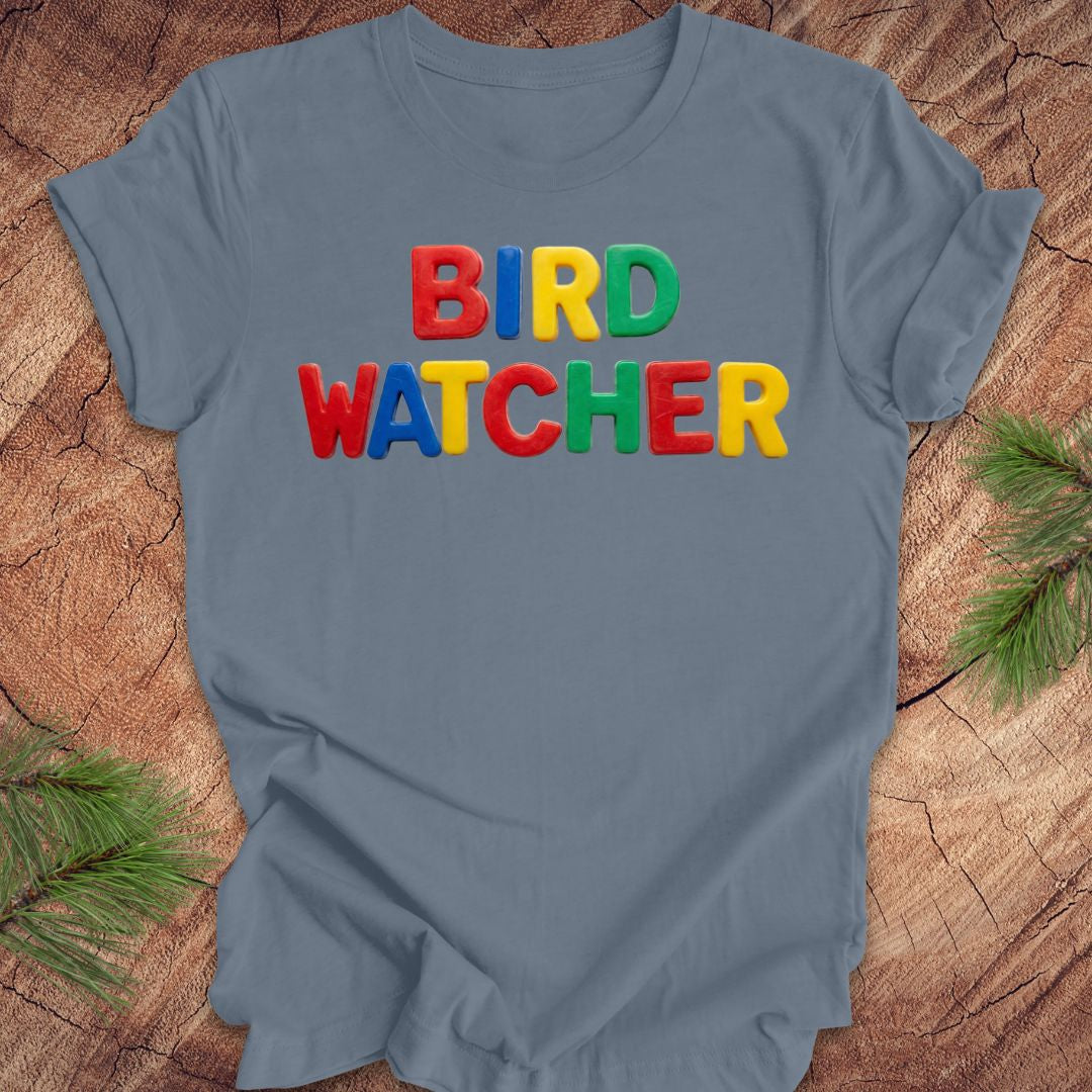Blue t-shirt with 'BIRD WATCHER' in colorful letters on a wooden surface.