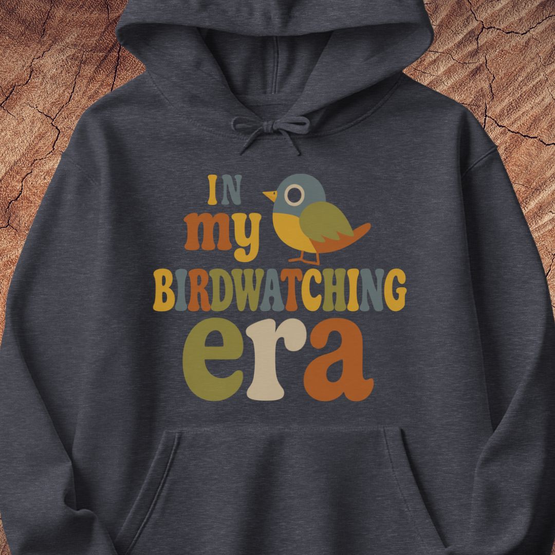 Dark heather gray hoodie with groovy retro IN MY BIRDWATCHING ERA lettering and a little songbird, laid flat on a rustic wood background.