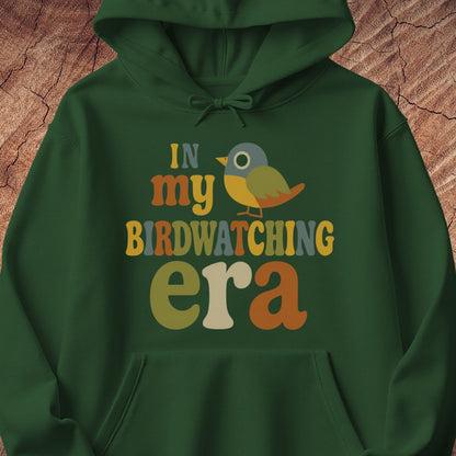 Forest green hoodie with groovy retro IN MY BIRDWATCHING ERA lettering and a little songbird, laid flat on a rustic wood background.