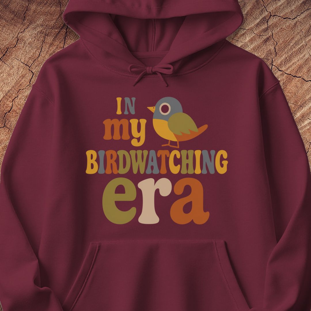 Maroon hoodie with groovy retro IN MY BIRDWATCHING ERA lettering and a little songbird, laid flat on a rustic wood background.