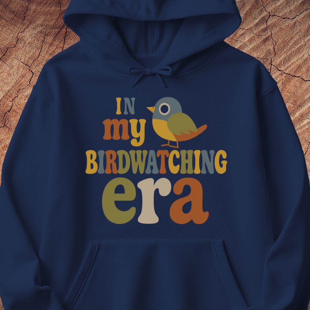 Navy hoodie with groovy retro IN MY BIRDWATCHING ERA lettering and a little songbird, laid flat on a rustic wood background.