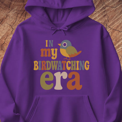 Purple hoodie with groovy retro IN MY BIRDWATCHING ERA lettering and a little songbird, laid flat on a rustic wood background.