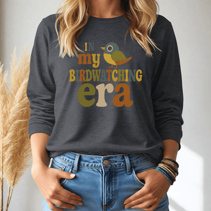 Person wearing a dark gray long-sleeve tshirt with colorful text 'In my birdwatching era' and a bird graphic, standing against a neutral background.