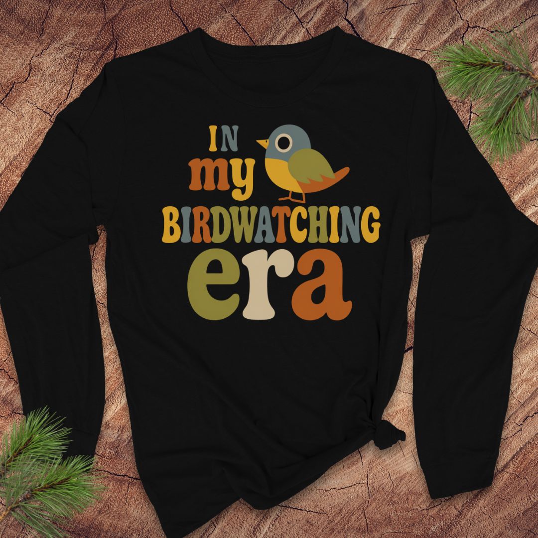 A black long-sleeve t-shirt with a colorful graphic that reads 'IN my BIRDWATCHING era' with a small bird illustration, laid flat on a wood surface with pine branches in the corners.