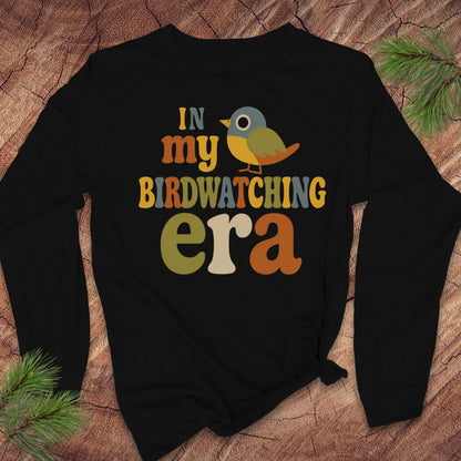 A black long-sleeve t-shirt with a colorful graphic that reads 'IN my BIRDWATCHING era' with a small bird illustration, laid flat on a wood surface with pine branches in the corners.