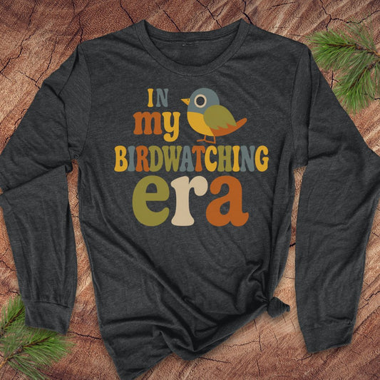 A dark charcoal gray long-sleeve t-shirt with a colorful graphic reading 'IN my BIRDWATCHING era' and a small bird illustration, laid flat on a wood surface with pine branches in the corners.