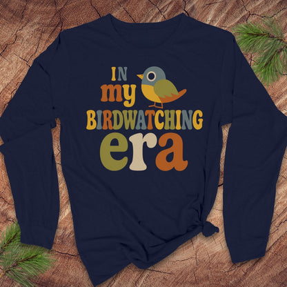 A navy blue long-sleeve t-shirt with a colorful graphic reading 'IN my BIRDWATCHING era' and a small bird illustration, laid flat on a wood surface with pine branches in the corners.