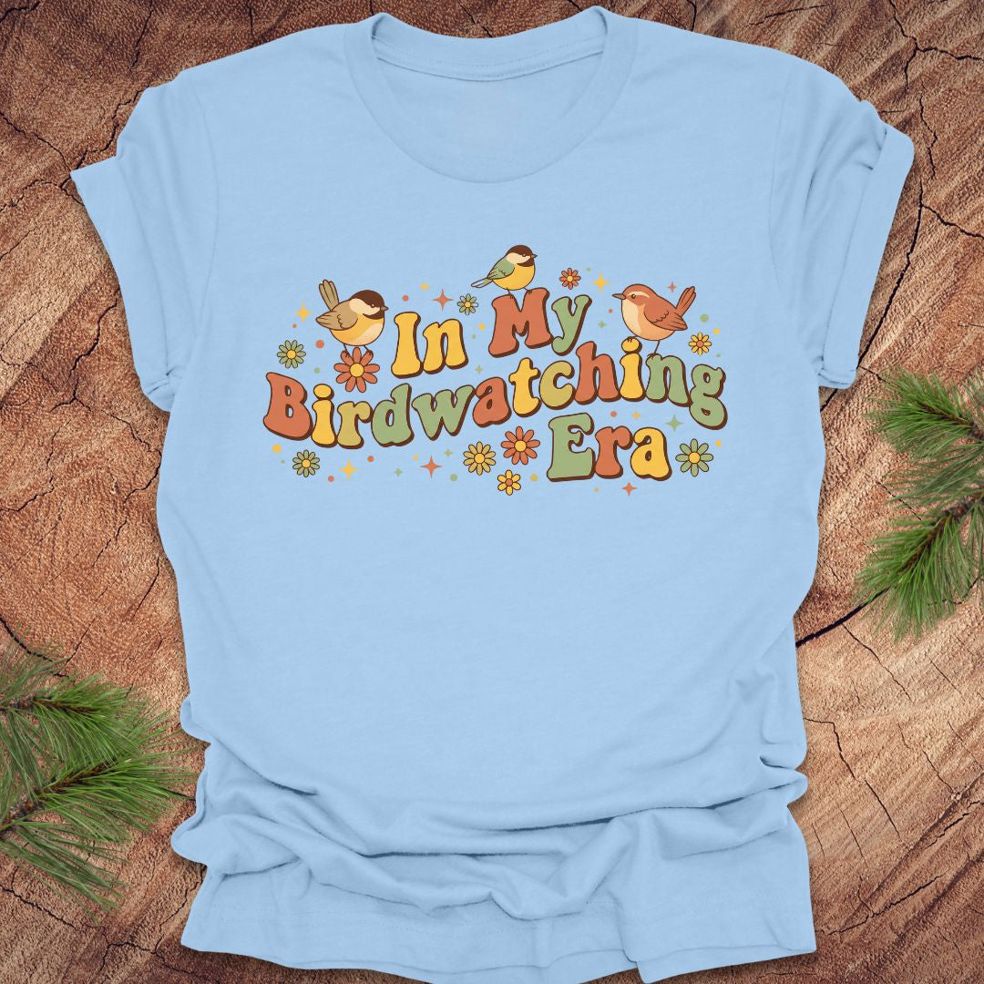 Light blue t-shirt with 'In My Birdwatching Era' text and bird illustrations on a wooden surface.