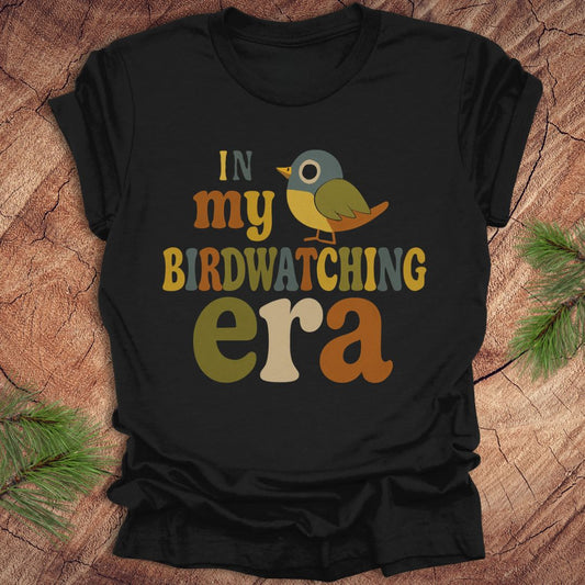 Black t-shirt with a colorful bird graphic and text, 'in my birdwatching era' on a wooden surface with pine branches.