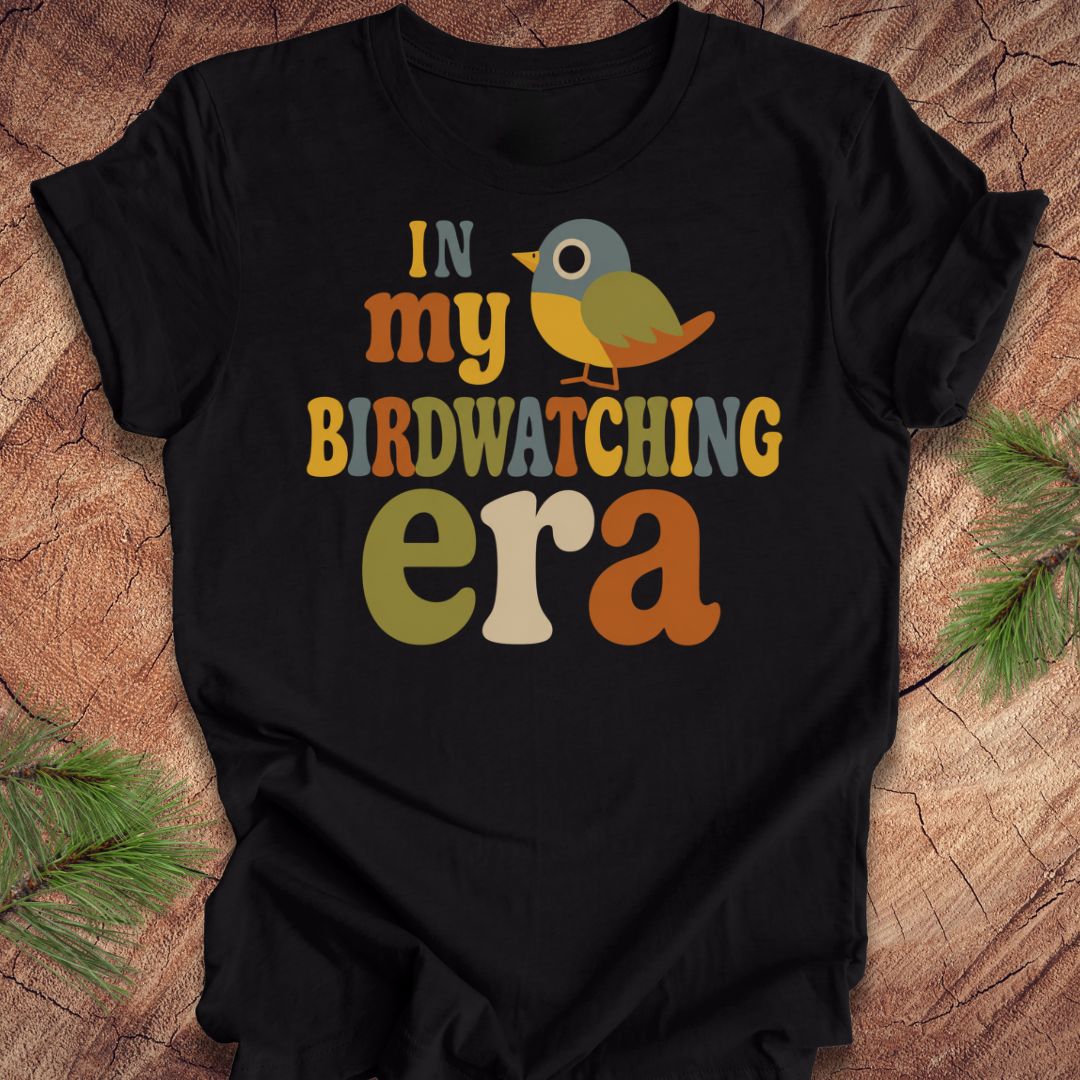 A black short-sleeve t-shirt with a colorful retro graphic that reads 'IN my BIRDWATCHING era' with a small bird illustration, laid flat on a rustic wood background with pine branches in the corners.
