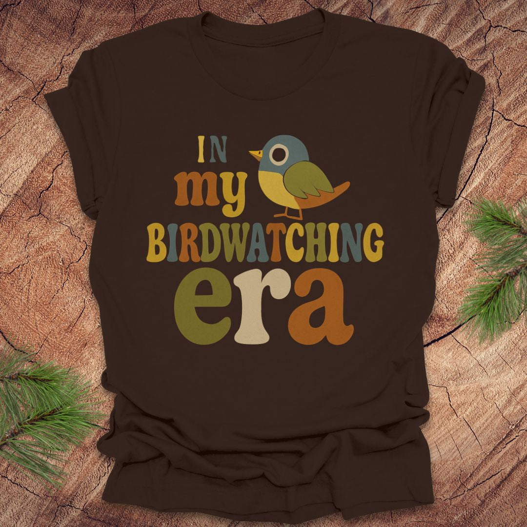 Brown t-shirt with a colorful bird graphic and text, 'in my birdwatching era' on a wooden surface with pine branches.