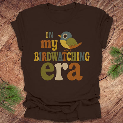 Brown t-shirt with a colorful bird graphic and text, 'in my birdwatching era' on a wooden surface with pine branches.