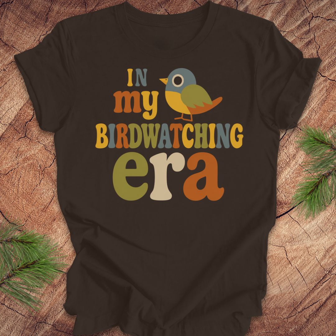 A dark brown graphic t-shirt laid flat on a rustic wood background with pine branches in the corners. The shirt features a colorful retro-style graphic that reads 'IN my BIRDWATCHING era' with a small bird illustration.