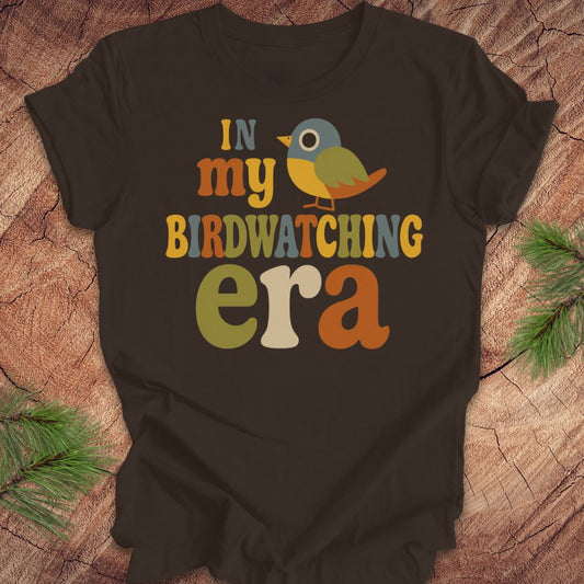 A dark brown graphic t-shirt laid flat on a rustic wood background with pine branches in the corners. The shirt features a colorful retro-style graphic that reads 'IN my BIRDWATCHING era' with a small bird illustration.