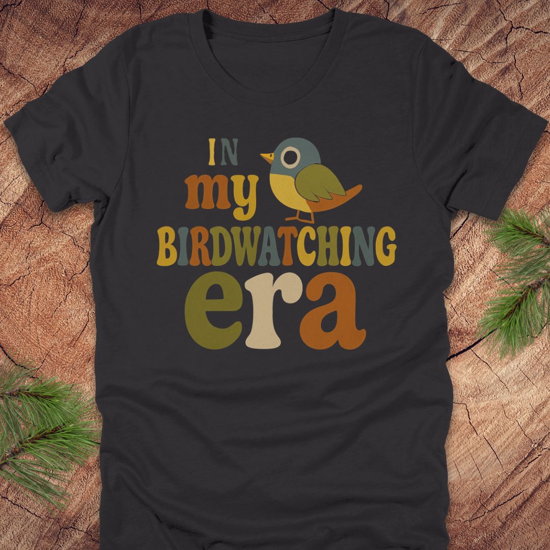 Dark grey t-shirt with a colorful bird graphic and text, 'in my birdwatching era' on a wooden surface with pine branches.