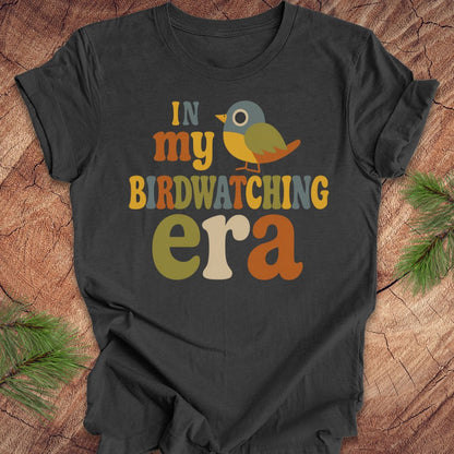 A dark charcoal gray t-shirt laid flat on a rustic wood background with pine branches in the corners. The shirt features a colorful graphic that reads 'IN my BIRDWATCHING era' with a small stylized bird in the center.