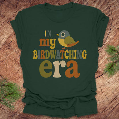 Green t-shirt with a colorful bird graphic and text, 'in my birdwatching era' on a wooden surface with pine branches.
