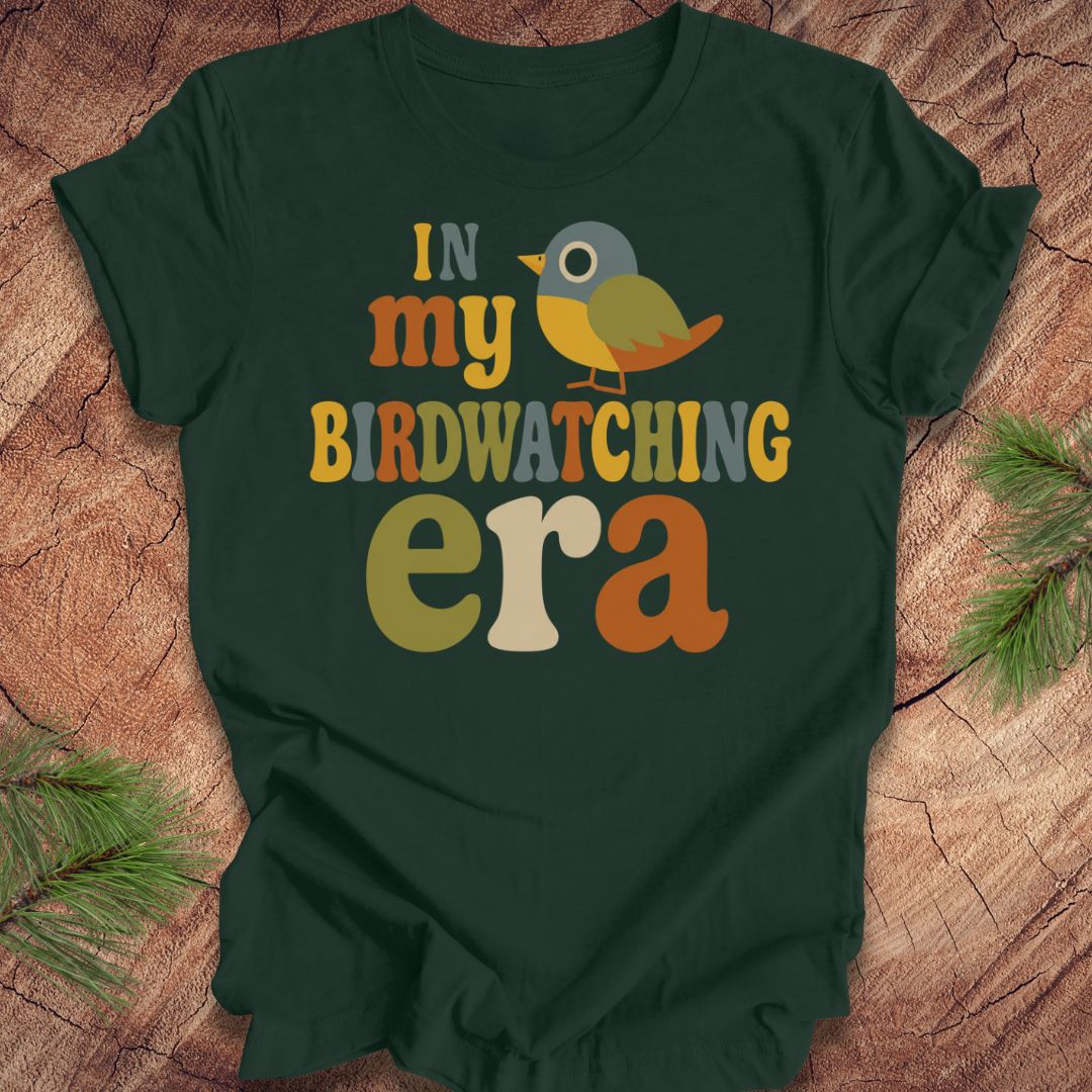 A dark forest green t-shirt with a colorful retro-style graphic reading 'IN my BIRDWATCHING era' and a small bird illustration, laid flat on a wood surface with pine branches in the corners.
