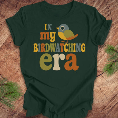 A dark forest green t-shirt with a colorful retro-style graphic reading 'IN my BIRDWATCHING era' and a small bird illustration, laid flat on a wood surface with pine branches in the corners.