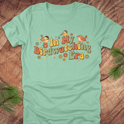 Mint green t-shirt with 'In My Birdwatching Era' text and birds on a wooden surface.