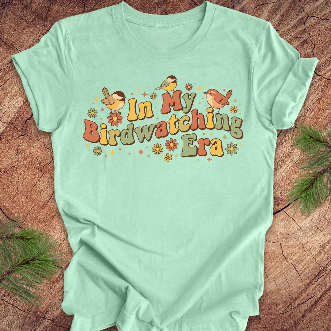 Mint green t-shirt with 'In My Birdwatching Era' text and birds on a wooden surface.