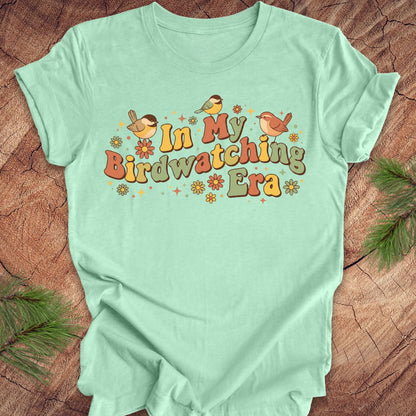 Mint green t-shirt with 'In My Birdwatching Era' text and birds on a wooden surface.