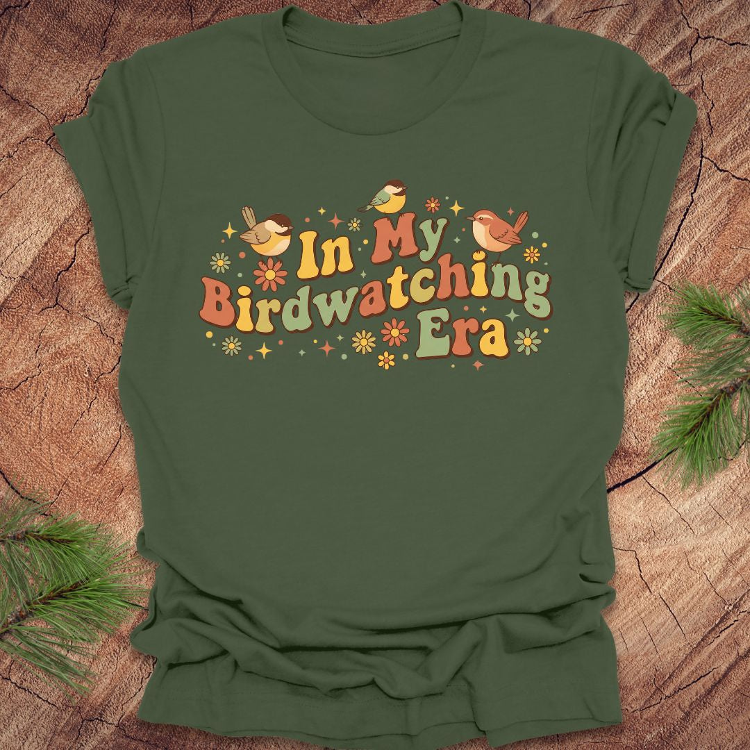 Green t-shirt with 'In My Birdwatching Era' text and bird illustrations on a wooden surface.