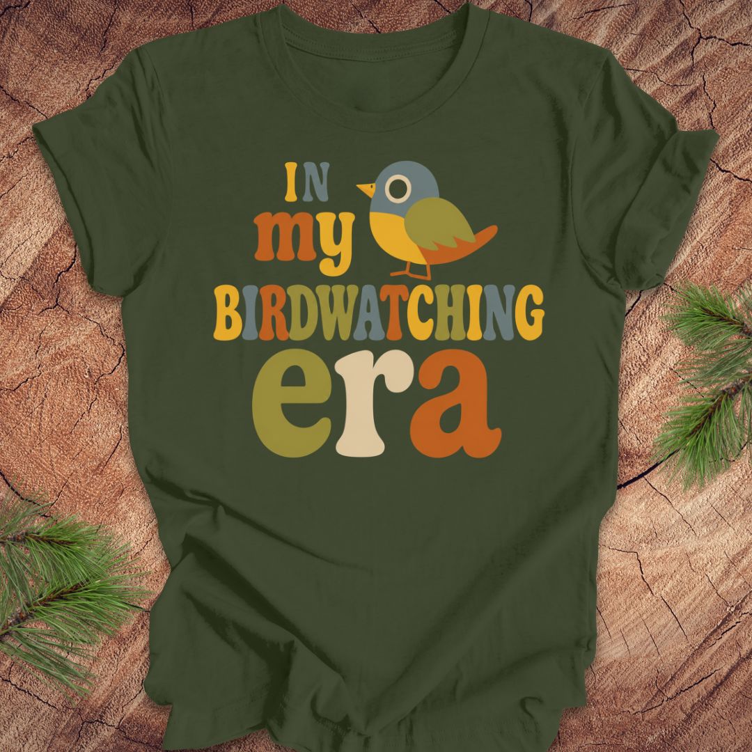 An olive green short-sleeve t-shirt with a colorful retro graphic that reads 'IN my BIRDWATCHING era' with a small bird illustration, laid flat on a rustic wood background with pine branches.