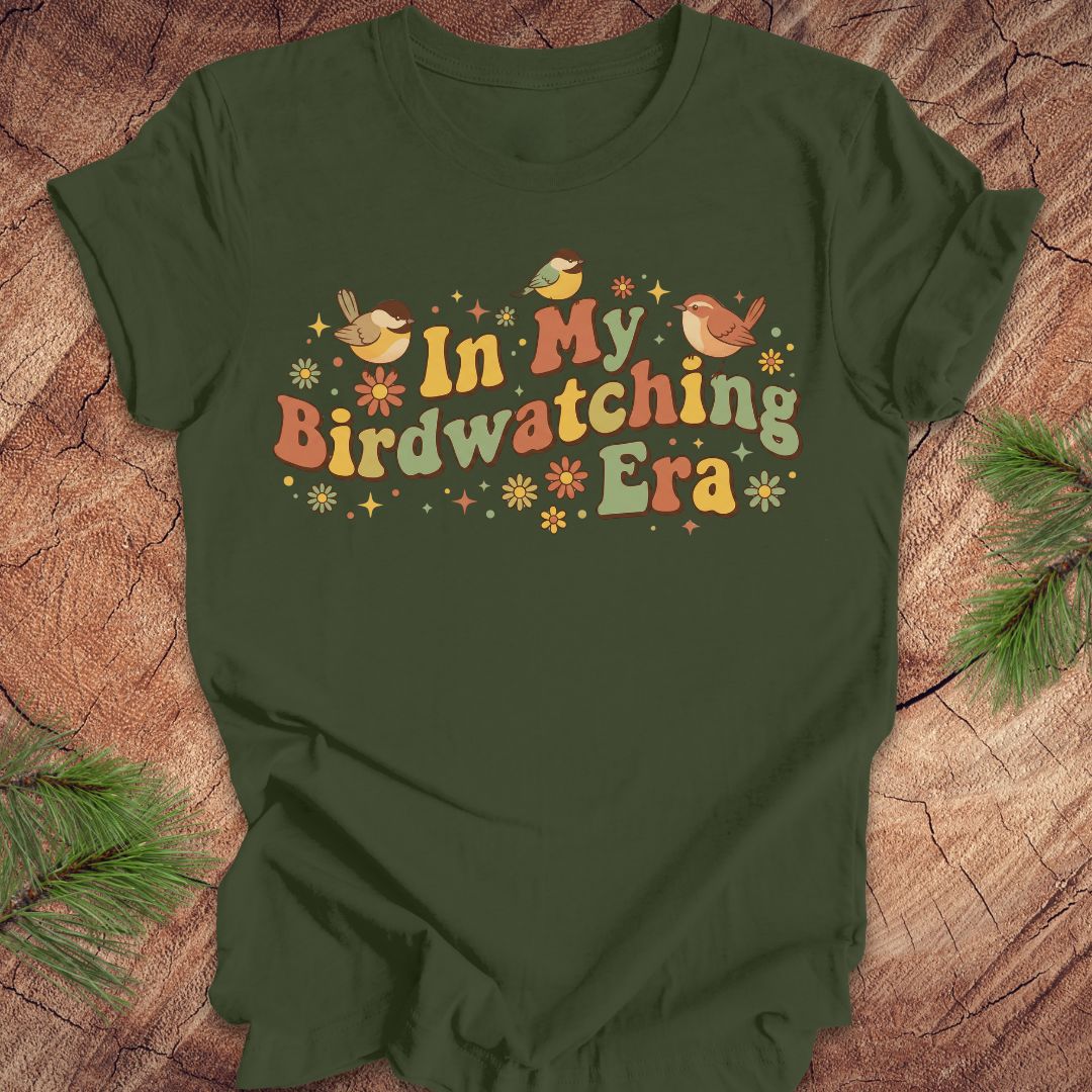 Green t-shirt with 'In My Birdwatching Era' text and bird illustrations on a wooden surface.