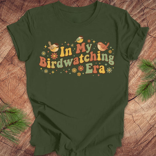 Green t-shirt with 'In My Birdwatching Era' text and bird illustrations on a wooden surface.