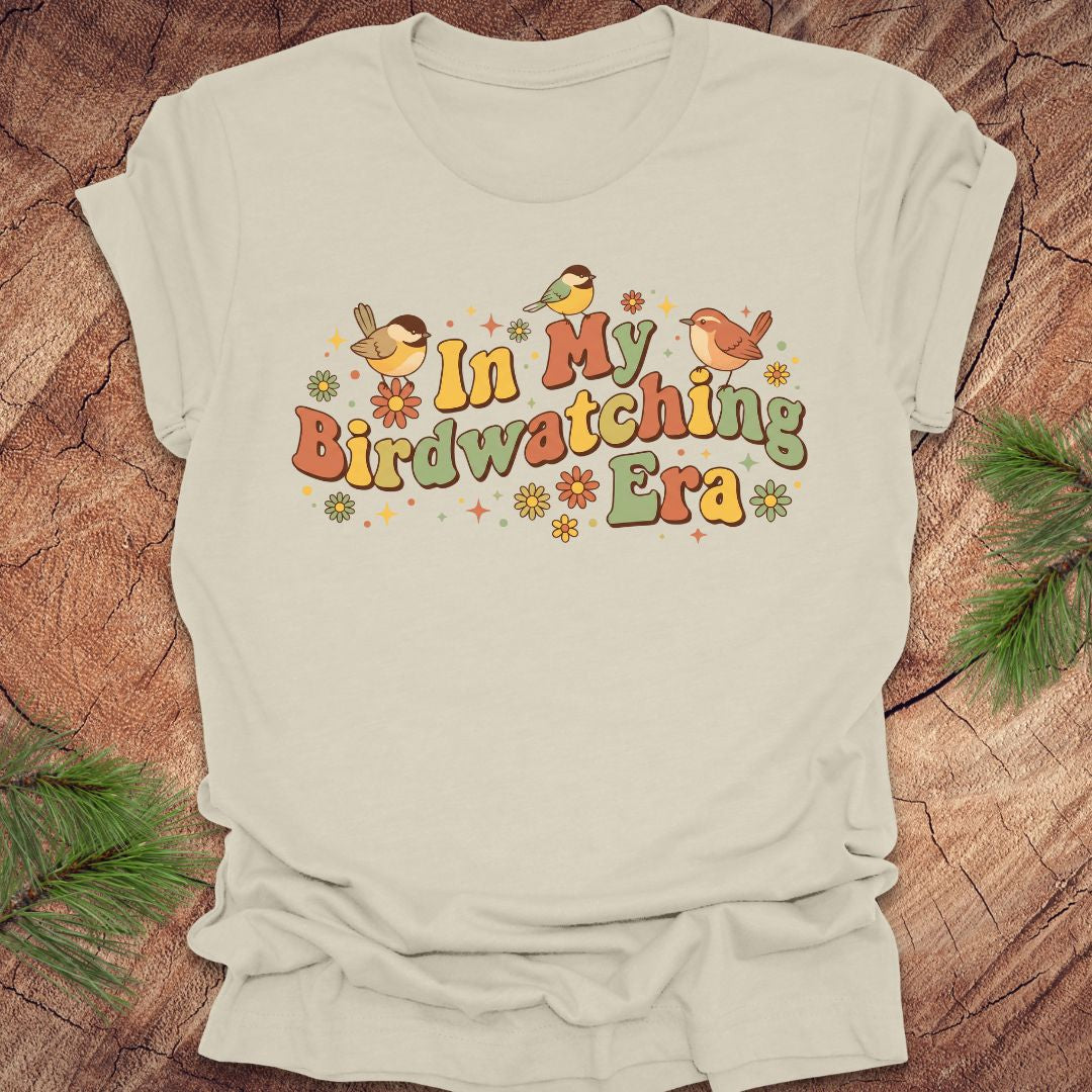 Beige t-shirt with 'In My Birdwatching Era' text and bird illustrations on a wooden surface.