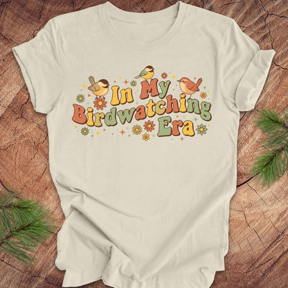 Beige t-shirt with 'In My Birdwatching Era' text and bird illustrations on a wooden surface.