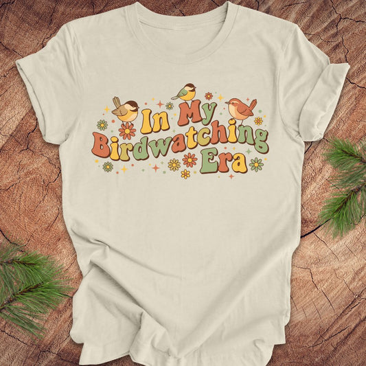 Beige t-shirt with 'In My Birdwatching Era' text and bird illustrations on a wooden surface.
