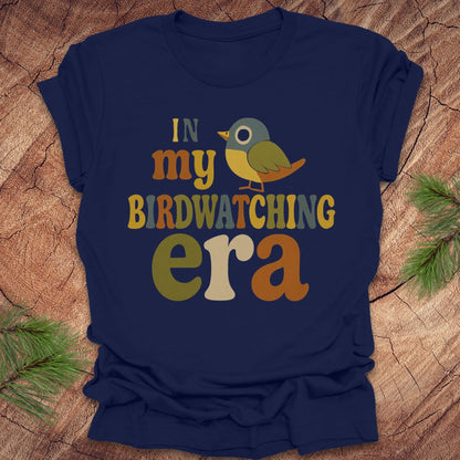 Navy blue t-shirt with a colorful bird graphic and text, 'in my birdwatching era' on a wooden surface with pine branches.