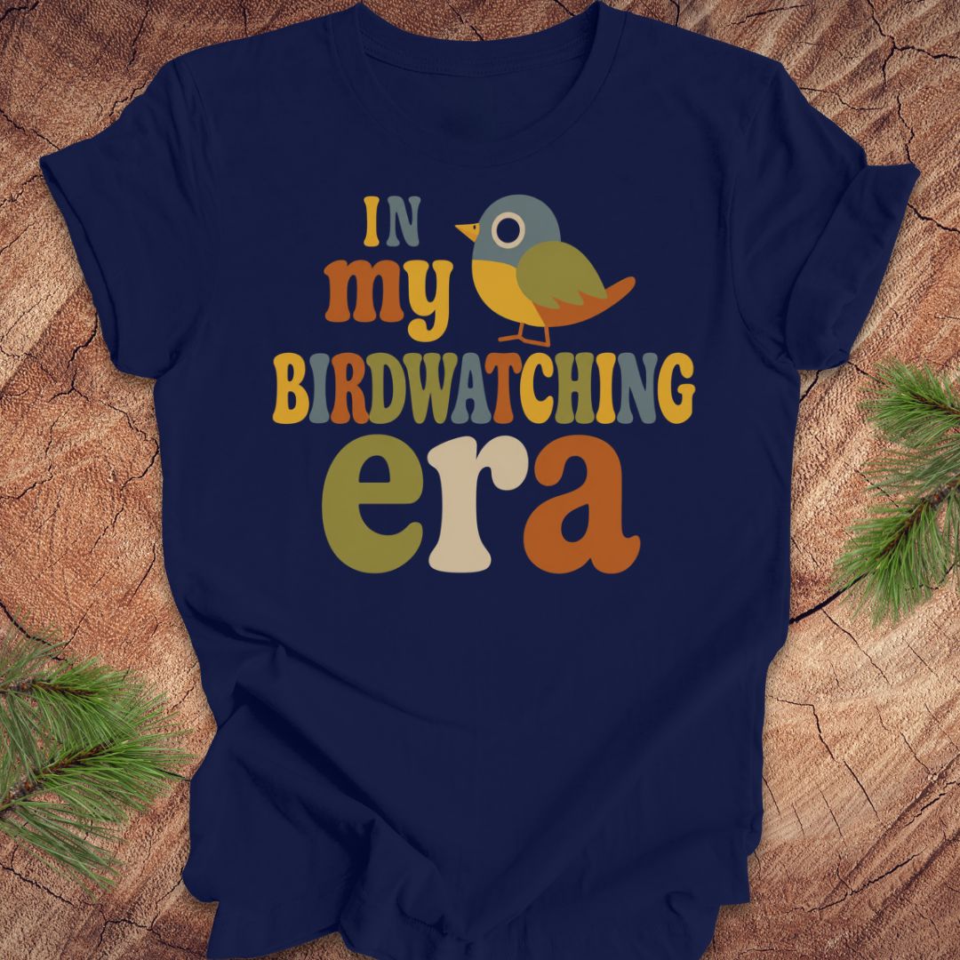 A navy blue short-sleeve t-shirt with a colorful graphic reading 'IN my BIRDWATCHING era' and a small bird illustration, laid flat on a wood surface with pine branches in the corners.