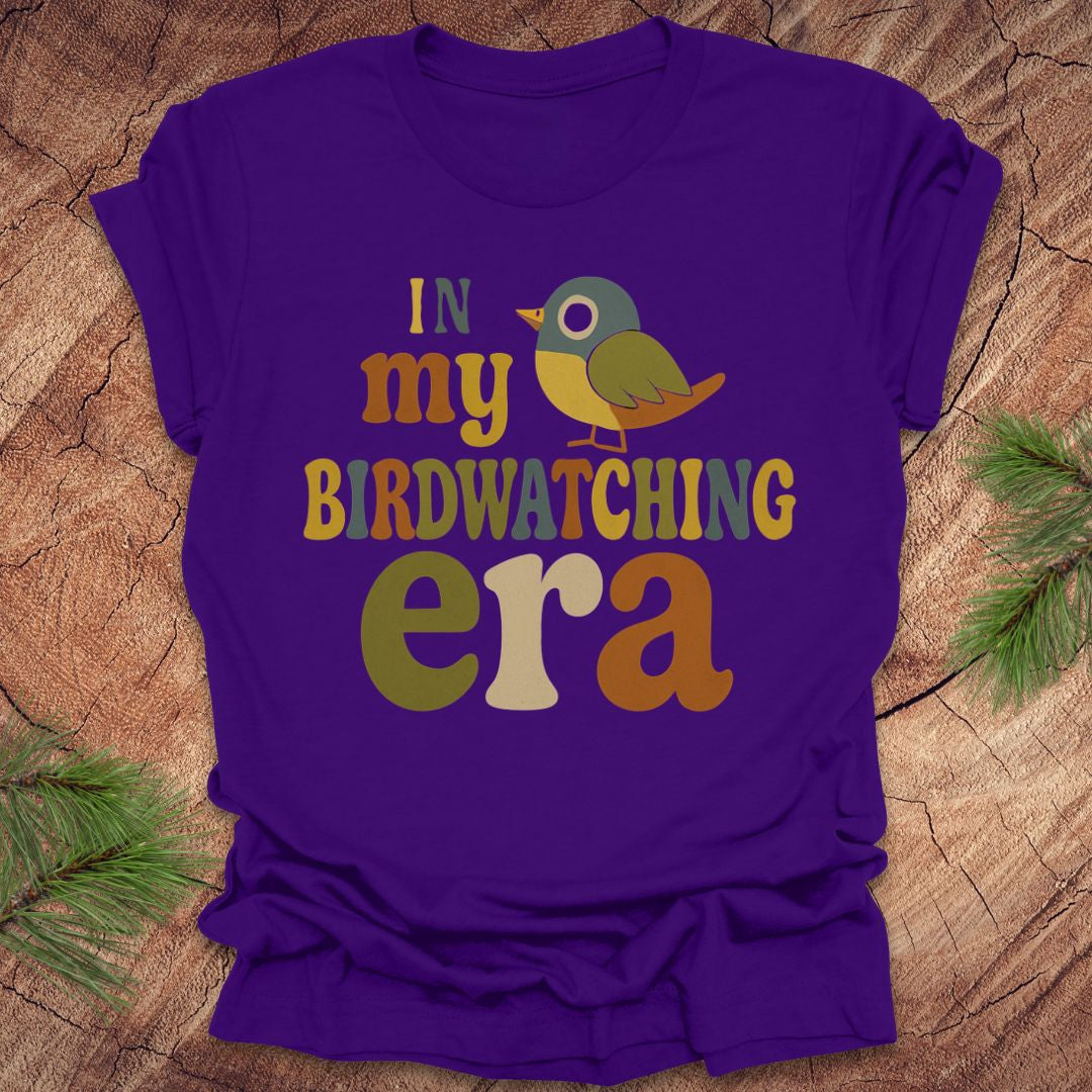 Purple t-shirt with a colorful bird graphic and text, 'in my birdwatching era' on a wooden surface with pine branches.