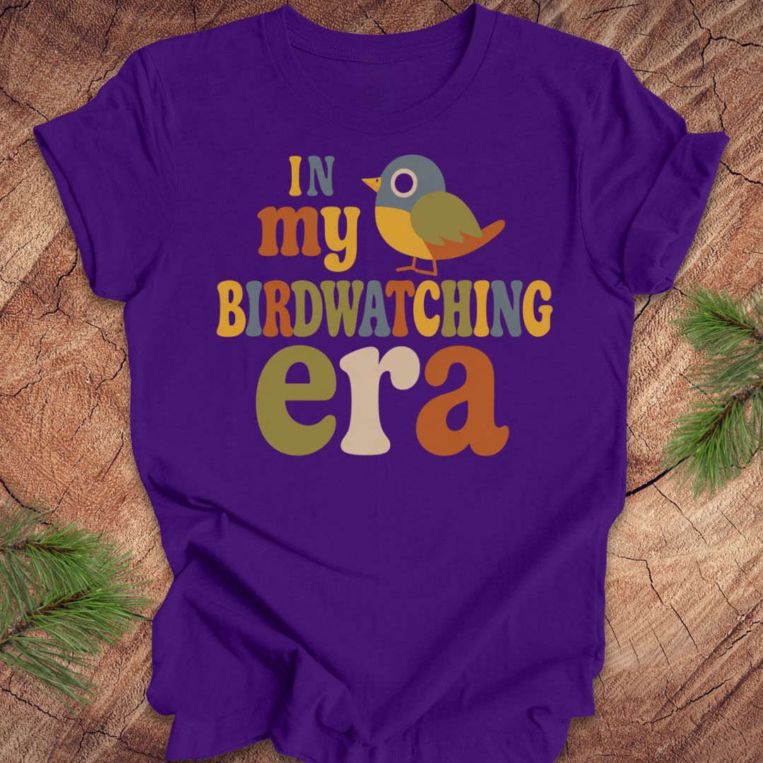 A purple short-sleeve t-shirt with a colorful graphic reading 'IN my BIRDWATCHING era' and a small bird illustration, laid flat on a wood surface with pine branches around it.