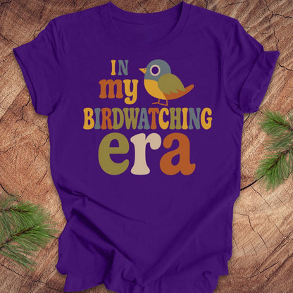 A purple short-sleeve t-shirt with a colorful graphic reading 'IN my BIRDWATCHING era' and a small bird illustration, laid flat on a wood surface with pine branches around it.