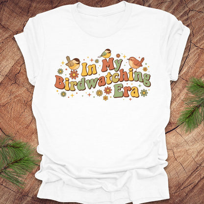 White t-shirt with 'In My Birdwatching Era' text and bird illustrations on a wooden surface.