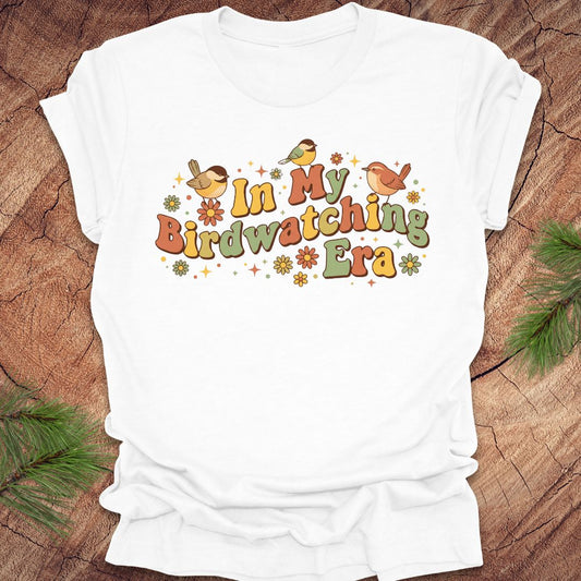 White t-shirt with 'In My Birdwatching Era' text and bird illustrations on a wooden surface.